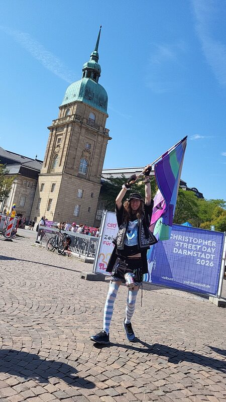 Person mit zwei Fahnen vor dem Turm am Karolinenplatz, daneben das CSD-Banner